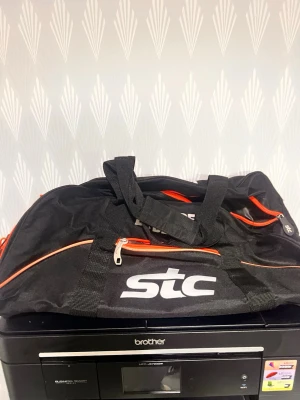 Svart sportbag från STC - Säljer en svart sportbag från STC med vita logotyper och texten 'DO MORE BE BETTER'. Väskan har flera fack med orange dragkedjor och bärhandtag. Perfekt för gymmet eller träning. Tillverkad i slitstarkt tygmaterial.
