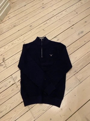 Mörkblå halvzip tröja från Gant - Mörkblå stickad tröja från Gant med halv dragkedja och liten logga på bröstet. Tröjan har ribbade muddar och en hög krage. Perfekt för lager på lager och har en snygg struktur i materialet.