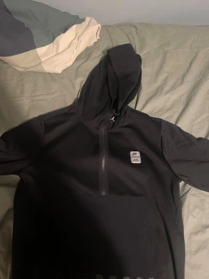 Svart Nike Air Max hoodie med huva - Snygg svart hoodie från Nike Air Max med stor magficka och halv dragkedja framtill. Har en tydlig AIR MAX-logga på fickan och en patch med Nike-logga på bröstet. Tillverkad i mjukt material och har en klassisk huva för extra stilpoäng.