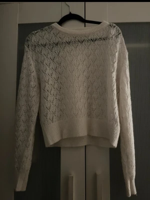 Vit spetsstickad tröja från H&M - Säljer en vit, spetsstickad tröja från H&M med lång ärm och ribbade muddar. Tröjan har ett fint hålmönster och är lite kortare i modellen, perfekt för att skapa en trendig lager-på-lager-look.