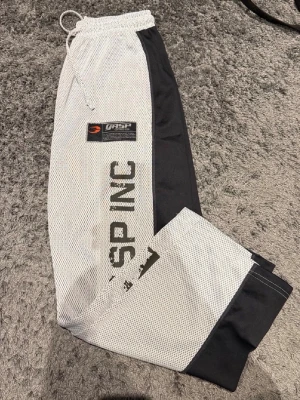 Svarta och vita meshbyxor från GASP - Säljer ett par sportiga träningsbyxor från GASP i svart och vitt meshmaterial. Byxorna har resår och dragsko i midjan, tryckt logga och texten GASP INC på benet. Perfekta för gymmet eller annan träning. Använda 3 gånger alltså i nyskick