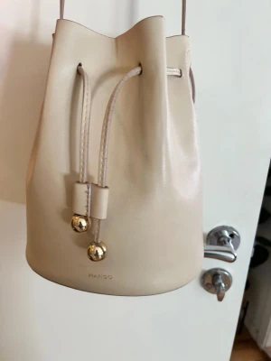 Beige bucket bag från Mango - Snygg beige bucket bag från Mango i slätt konstläder. Väskan har dragskostängning med guldfärgade detaljer och en smal axelrem. Perfekt för dig som gillar minimalistisk och stilren design.