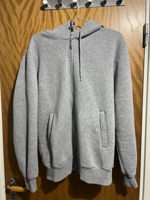 Grå zip-hoodie med fickor - En klassisk grå hoodie med dragkedja framtill och två sidofickor. Tröjan har huva med dragsko och ribbade muddar vid ärmslut och nederkant. Perfekt för en chill och avslappnad stil. Materialet känns mjukt och bekvämt, troligen bomullsblandning.