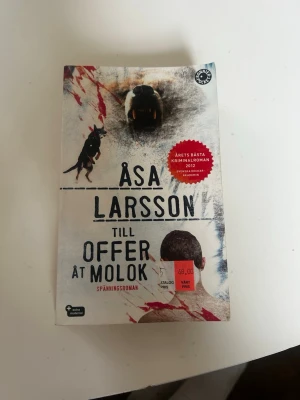 Till offer åt Molok av Åsa Larsson - Spännande kriminalroman av Åsa Larsson med omslag i vitt och blodröda detaljer. Boken är en pocketutgåva och har ett omslag med björn, hund och blodstänk. Perfekt för dig som gillar nervkittlande deckare och nordisk spänning.