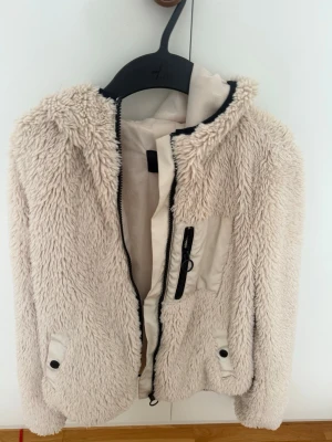 Beige teddyjacka från Vero Moda - Mysig beige teddyjacka från Vero Moda med huva och dragkedja framtill. Jackan har två fickor med knappstängning och en bröstficka med dragkedja. Perfekt för kalla dagar när du vill vara både varm och stylish.
