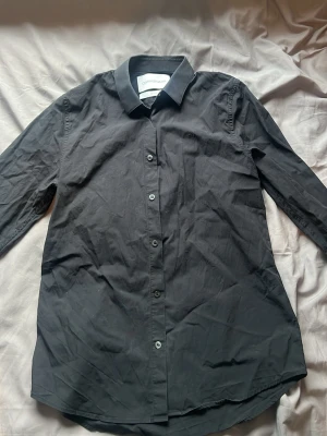Svart skjorta från Calvin Klein - Stilren svart skjorta från Calvin Klein Jeans med diskret CKJ-brodyr på bröstet. Klassisk krage, knäppning framtill och lång ärm. Skjortan är i mjuk bomull och har en rak passform som funkar till många olika looks.