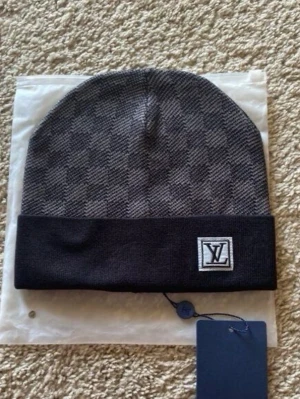 Grå mössa med logga - Säljer en snygg gråmelerad mössa med klassiskt rutmönster och vikt kant. På kanten sitter en stilren -logga i svart och vitt. Perfekt accessoar för dig som vill ha en exklusiv och trendig look. Materialet känns mjukt och bekvämt.