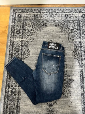 Blå slitna jeans från Born In Italy - Säljer ett par blå jeans från Born In Italy med slitningar och distressed detaljer. Jeansen har en smal passform, klassiska fem fickor och coola slitna partier samt färgstänk för en trendig look. Perfekta för dig som gillar streetstyle.