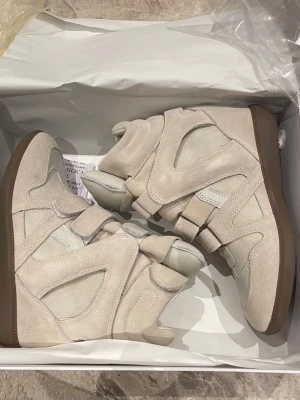 Isabel Marant skor - Säljer ett par ikoniska höga sneakers från Isabel Marant i vitt och beige mocka och läder. Skorna har chunky design, kardborreband och gummisula. Perfekta för dig som vill ha en statement sneaker med cool siluett och exklusiv känsla.