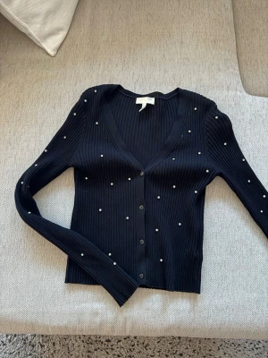 Svart ribbad kofta med pärlor H&M - Snygg svart ribbad kofta från H&M med dekorativa små pärlor över hela plagget. Koftan har v-ringning, långa ärmar och knäppning framtill. Perfekt för dig som gillar en stilren men ändå trendig look.