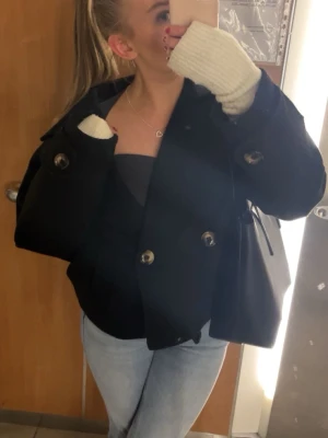 Svart dubbelknäppt kappa - Stilren svart kappa med dubbelknäppning och stora knappar. Klassisk krage och rak passform, perfekt för en trendig look. 