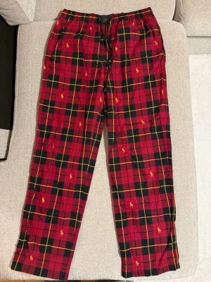 Rödrutiga pyjamasbyxor Polo Ralph Lauren - Snygga rödrutiga pyjamasbyxor från Polo Ralph Lauren med klassiskt rutmönster i rött, svart och gult. Byxorna har resår i midjan och små broderade loggor i gult. Tillverkade i mjuk bomull, perfekta för chillkvällar hemma.