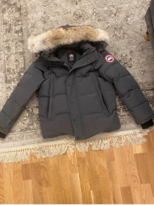Grå dunjacka från Canada Goose - Säljer en grå dunjacka från Canada Goose med klassisk patch på ärmen och huva. Jackan har en rak passform, dragkedja och knappar framtill samt ribbade muddar vid ärmsluten. Perfekt för kalla vinterdagar och riktigt varm tack vare dunfyllningen.
