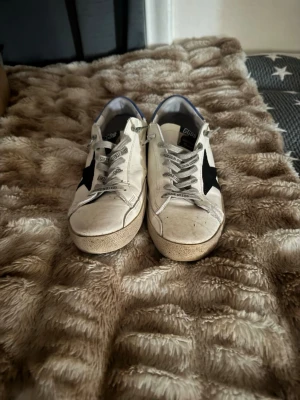 Golden Goose vita sneakers med blå detalj - Golden Goose sneakers i vitt läder med svart stjärna på sidan och blå hälkappa. Klassisk låg modell med grå skosnören och slitna detaljer för en cool, avslappnad look. Perfekta för dig som gillar streetstyle och unika skor.