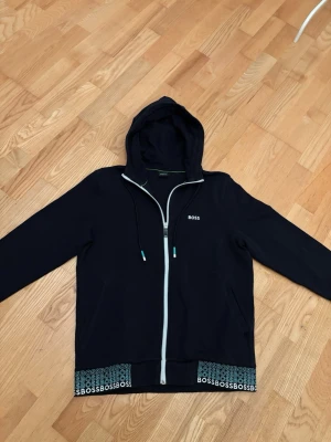 Svart zip-hoodie från BOSS - Svart hoodie med dragkedja från BOSS. Snygga detaljer med vit dragkedja och logga på bröstet samt BOSS-tryck längs nederkanten. Huva med dragsko och ribbade muddar. Perfekt för en chill och stilren look.