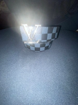 Rutmönstrat bällte  - Snyggt bälte med klassiskt rutmönster i svart och blått. Bältet har ett stort spänne i silverfärg och är tillverkat i skinn. Perfekt accessoar för att lyfta din outfit.