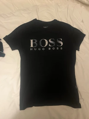 Svart Hugo Boss t-shirt med logga - Snygg svart t-shirt från Hugo Boss med stor vit BOSS-logga framtill. Klassisk rund halsringning och korta ärmar. Regular fit och tillverkad i mjuk bomull som känns skön mot huden. Perfekt för dig som gillar stilrena och ikoniska plagg.