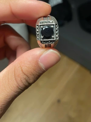 Silverfärgad ring med svart sten - Snygg silverfärgad ring med en stor svart sten i mitten och detaljerade mönster runt om. Ringen har en fyrkantig front med små kuldetaljer och graverade sidor. Perfekt för dig som gillar statement-smycken med en unik touch.