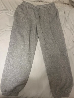 Gråa mjukisbyxor  - Säljer ett par klassiska gråa mjukisbyxor med resår i midjan och dragsko för perfekt passform. Byxorna har muddar vid bensluten och är gjorda i mjukt bomullsmaterial. Perfekta för chill dagar eller när du vill vara extra bekväm.