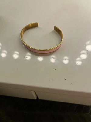 Guldigt armband med rosa detalj - Snyggt och stilrent armband i metall med guldfärgad finish och en ljusrosa emaljerad rand längs utsidan. Öppen design som gör det enkelt att ta på och av. Perfekt för dig som gillar minimalistiska accessoarer med en touch av färg.