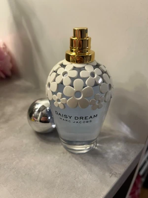 Daisy Dream parfym från Marc Jacobs - Daisy Dream från Marc Jacobs är en parfym i en ljusblå glasflaska med vita blommor i plast runt halsen och en guldfärgad spray. Designen är lekfull och feminin, perfekt för dig som gillar snygga detaljer💗Doften är blommig och fräsch med inslag av citrus och fruktiga toner🌸🍋Parfymen har endast testats en gång men förpackningen som den är kom i är tyvärr slängd  Öppen för bud💕