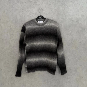 Études Knit - Säljer en stickad tröja från Études i gråmelerad färg med mjuk, luddig textur. Tröjan har rund halsringning och långa ärmar. Färgskiftningarna går i olika nyanser av grått och svart för en cool, modern look.