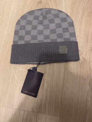 Zara - Säljer en snygg grå mössa från Vuitton med klassiskt rutmönster och vikt kant. Mössan har en diskret L-logga på en patch framtill och är tillverkad i mjukt, stickat material. Perfekt accessoar för dig som gillar exklusiv och stilren design.