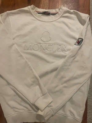 Vit sweatshirt från Moncler - Snygg vit sweatshirt från Moncler med broderad logga på bröstet och ett Moncler-märke på ärmen. Tröjan har rund halsringning, ribbade muddar och är tillverkad i mjukt bomullsmaterial. Perfekt för en clean och stilren look.