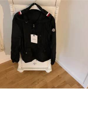 Svart Moncler vindjacka med huva - FAST PRIS.Snygg svart vindjacka från Moncler med huva och dragkedja framtill. Jackan har Moncler-logga på ärmen och detaljer i rött, vitt och blått på huvan och ärmsluten. Tillverkad i lätt och tåligt material, perfekt för blåsiga dagar.