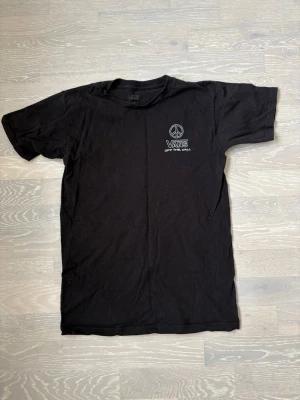 Svart Vans t-shirt med tryck - Svart t-shirt från Vans med ett vitt tryck på bröstet som visar ett peace-märke och texten 'Vans Off The Wall'. Klassisk passform och rund halsringning. Perfekt för dig som gillar streetwear och enkel stil.