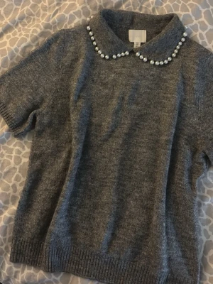 Grå stickad topp med pärlkrage - Säljer en grå stickad topp från H&M med korta ärmar och en söt krage dekorerad med pärlor.  Perfekt för dig som vill ha en trendig och unik detalj i garderoben.