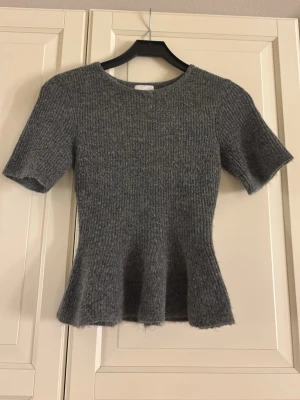 Grå stickad topp från H&M - Säljer en grå stickad topp från H&M med korta ärmar och figurnära passform. Toppen har en lätt utsvängd nederkant och rund halsringning. Perfekt för dig som gillar en enkel men snygg stil.