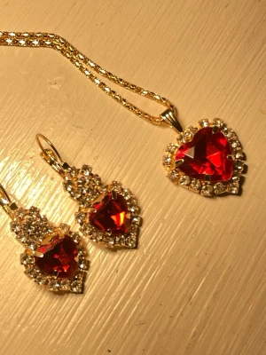 Rött hjärtsmyckeset med strass - Set med halsband och örhängen i guldtonad metall. Hjärtformade röda stenar omgivna av glittrande strass. Perfekt för att ge en glamorös touch till din outfit.