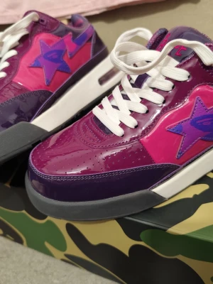 Verifierat äkta via checkcheck BAPE STA Sneakers EU 42,5 med original box och accessoarer - Ikoniska BAPE STA sneakers i den eftertraktade lila camo-designen, i storlek 42,5. Dessa exklusiva sneakers från det legendariska japanska streetwearmärket A Bathing Ape (BAPE) är helt nya och levereras med alla tillbehör, inklusive originalkartong och prisskylt. BAPE är känt för sin unika, högkvalitativa design som blandar streetwear och lyx, vilket gör varje produkt till ett samlarobjekt. Sneakers från BAPE är både modeikoniska och eftertraktade bland sneakerheads och modeentusiaster världen ö