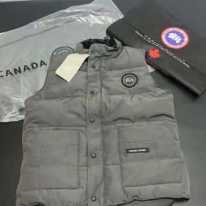 Grå dunväst  - Snygg grå dunväst med hög krage och tryckknappar framtill. Västen har två stora fickor med lock och en diskret logotyp på bröstet samt en patch på ena fickan. Perfekt för lager-på-lager och streetwear.