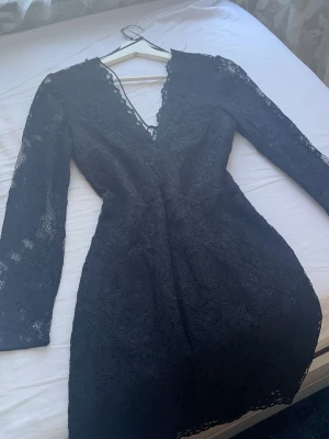 Svart lace klänning med v-ringning - Snygg svart kortklänning i spets med långärmad design och djup v-ringning. Klänningen har figurnära passform och fina spetsdetaljer längs kanterna. Perfekt för dig som vill ha en elegant och trendig look.
