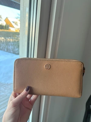 Beige/brun plånbok från Tory Burch - Stilren beige plånbok från Tory Burch i saffiano-läder med guldfärgad logga framtill. Plånboken har dragkedja runtom, flera kortfack och ett myntfack med dragkedja. Perfekt för att hålla ordning på kort och kontanter. Väldigt bra skick. Säljer för 1700kr