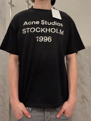 Svart Acne Studios t-shirt - Helt ny svart t-shirt från Acne Studios med vit text 'Acne Studios STOCKHOLM 1996' på bröstet. Tröjan är helt ny och lappar sitter kvar. Tröjan är i storlek S men den sitter relaxed så den passar M. Hör av er vid frågor 🙌