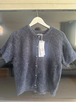 Mörkblå bouclékofta med kort ärm - Kortärmad kofta från Gina Tricot i mörkblå bouclé med rund hals och små guldfärgade knappar framtill. Koftan har en mjuk, fluffig textur och en avslappnad passform som ger en trendig look. Perfekt att styla över ett linne eller t-shirt. Helt ny oanvänd med prislapp. Ordinarie pris 399kr