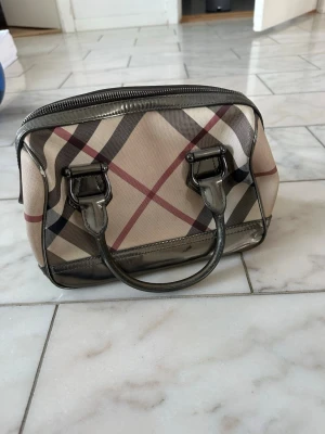 Beige handväska med rutigt mönster - Snygg beige handväska från Burberry med det klassiska rutiga mönstret i svart, rött och vitt. Väskan har metallicfärgade handtag och dragkedja, samt en innerficka med dragkedja. Tillverkad i canvas och läderdetaljer. Perfekt för dig som gillar tidlös design.