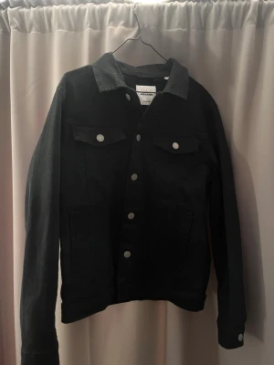 Svart jeansjacka från Jack & Jones - Snygg svart jeansjacka från Jack & Jones med klassisk krage, silverfärgade knappar och två bröstfickor med lock. Jackan har en rak passform och är tillverkad i slitstarkt jeansmaterial. Perfekt för dig som vill ha en stilren och tidlös look.
