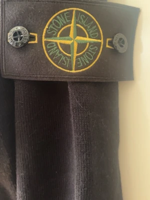 Stone Island Svart stickad tröja  - Snygg svart stickad tröja från Stone Island med den klassiska logotypen på ärmen. Tröjan har ribbade muddar och är tillverkad i mjukt material som ger en clean och stilren look. Perfekt för dig som gillar streetwear och vill ha något tidlöst.