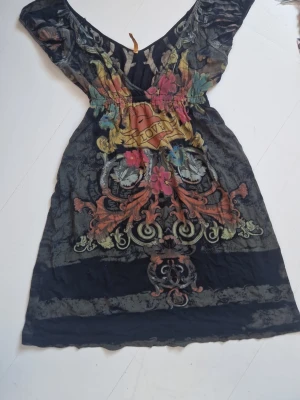 Svart kort klänning med färgglatt tryck - Unik svart kort babydoll-klänning med supersnyggt ed hardy-liknande färgglatt tryck och djup v-ringning, korta ärmar och resår i midjan för en snygg passform. 