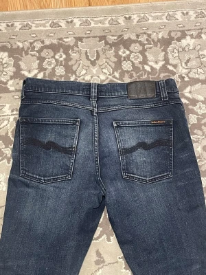 Nudie jeans Lean Dean  - Snygga mörkblå jeans från Nudie Jeans med klassisk femficksmodell och smal passform. Jeansen har kontrastsömmar, svarta broderade detaljer på bakfickorna och patch med logga i midjan. Tillverkade i mjuk denim med lätt tvättad look. Storlek 31/32