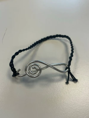 Armband med silverfärgad G-klav - Säljer ett armband gjort av flätad svart tråd. Fäst till svarta tråden är det en silverfärgad G-klav gjord av metalltråd. Perfekt för dig som vill ha ett smycke med en kreativ twist. 
