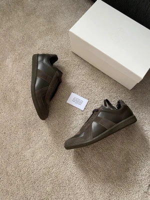 Maison Margiela Exklusiva Bruna Gats - Snygga sneakers i mörkbrunt läder och mocka från Maison Margiela. Klassisk low-top siluett med ton-i-ton detaljer och gummisula. Perfekta för dig som gillar minimalistisk och stilren design.