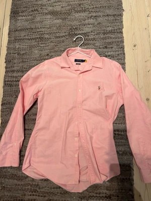 Rosa skjorta från Polo Ralph Lauren - Klassisk rosa skjorta från Polo Ralph Lauren med broderad logga på bröstet. Skjortan har lång ärm, knappar framtill och en snygg krage. Tillverkad i mjuk bomull för en bekväm känsla och stilren look. Storlek L men funkar för M. Knappt använd 