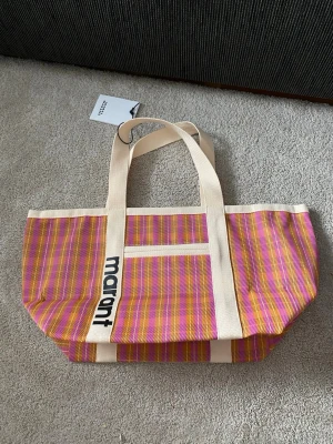 Randig Tote Bag från Isabel Marant - Färgglad shoppingväska från Isabel Marant med breda handtag och randigt mönster i rosa, orange och beige. Väskan har en stor öppen insida och är tillverkad i ett tåligt tygmaterial. Logotypen 'marant' syns tydligt på ena handtaget. Perfekt för att bära allt du behöver.