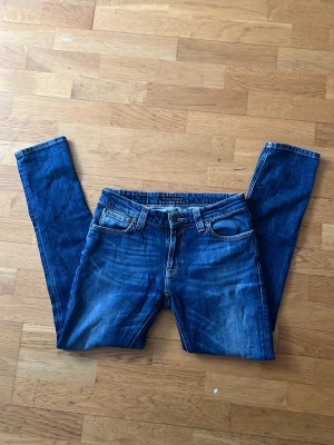 Blå skinny jeans från Nudie Jeans - Säljer ett par klassiska blå skinny jeans från Nudie Jeans med snygga slitningar och kontrastsömmar. Jeansen har fem fickor, bälteshällor och knappgylf. Perfekta för dig som gillar en smal passform och tidlös denimstil.