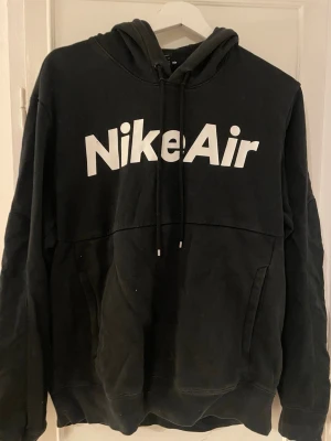 Svart Nike Air hoodie med huva - Svart hoodie från Nike med stor vit Nike Air-logga på bröstet. Klassisk huva med dragsnören och känguruficka framtill. Tillverkad i mjukt bomullsmaterial som är skönt att ha på sig. Perfekt för en avslappnad och sportig stil.